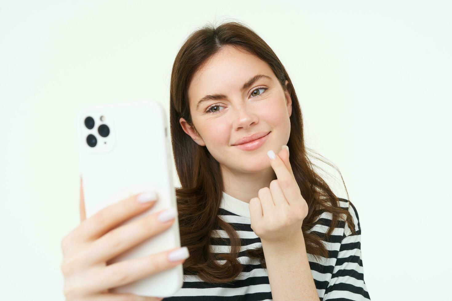 Porträt von schön Frau zeigen Herz Zeichen und posieren zum Selfie, nehmen Bild von Sie selber auf Smartphone Anwendung, posieren in der Nähe von etwas Niedlich, Weiß Hintergrund foto