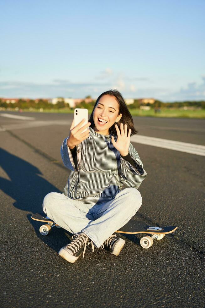 glücklich Teen asiatisch Mädchen nimmt Selfie, Video Chats auf Smartphone App während sitzt auf Skateboard, Skater Aufzeichnungen vlog während sitzt auf Longboard foto