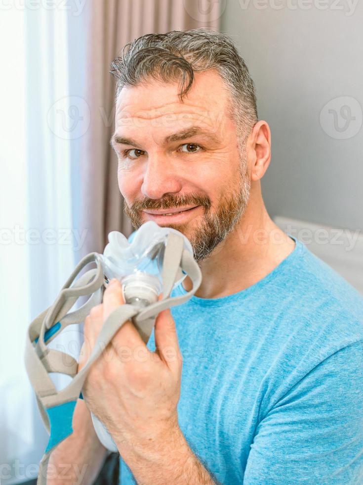 Reifer glücklicher Mann mit chronischen Atemproblemen erwägt, mit einer CPAP-Maschine auf dem Bett im Schlafzimmer zu sitzen. Gesundheitswesen, CPAP, Schnarchkonzept foto