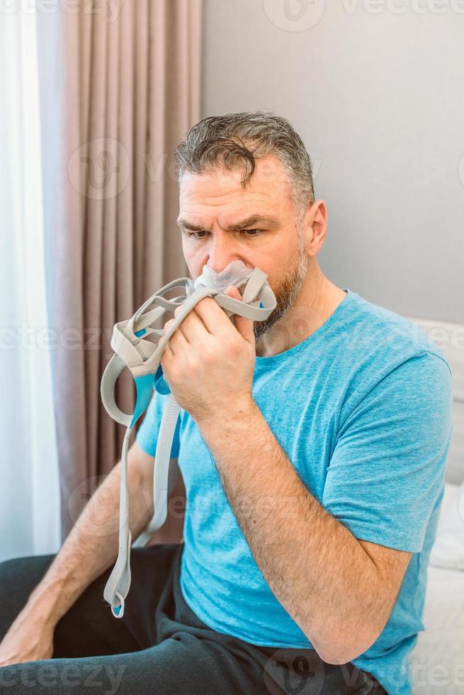 unglücklicher schockierter mann mit chronischen atmungsproblemen, überrascht von der verwendung der cpap-maschine, die auf dem bett im schlafzimmer sitzt. Gesundheitsversorgung, CPAP, obstruktive Schlafapnoe-Therapie, Schnarchkonzept foto