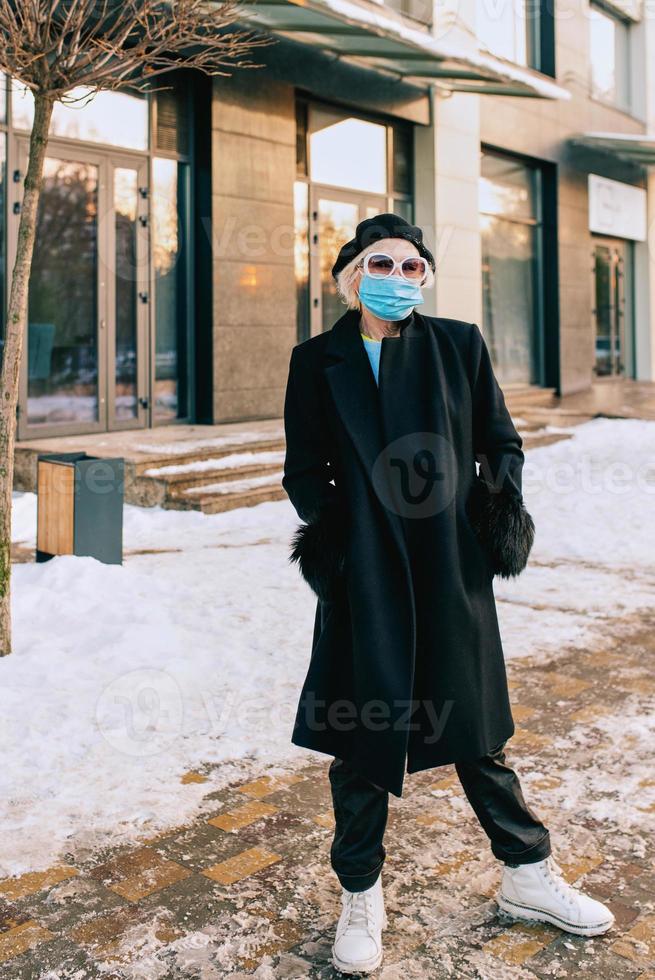 Senior stilvolle Frau in Baskenmütze und elegantem schwarzem Mantel und in medizinischer Maske, die im Freien geht. Lockdown, Pandemie, Schutzkonzept foto