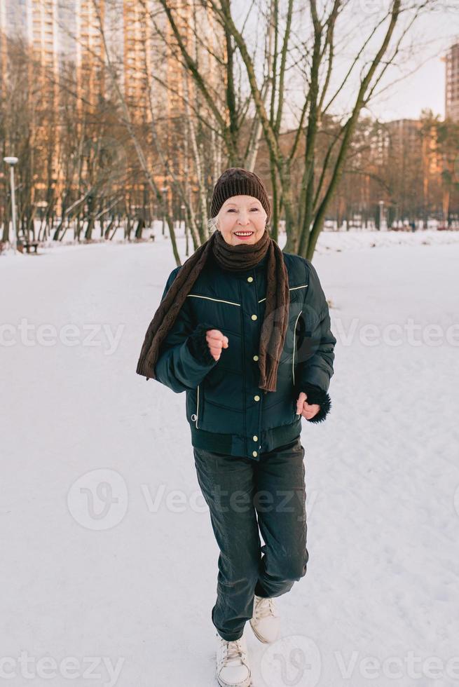 ältere Frau mit Hut und sportlicher Jacke beim Joggen im Schneewinterpark. Winter, Alter, Sport, Aktivität, Saisonkonzept foto