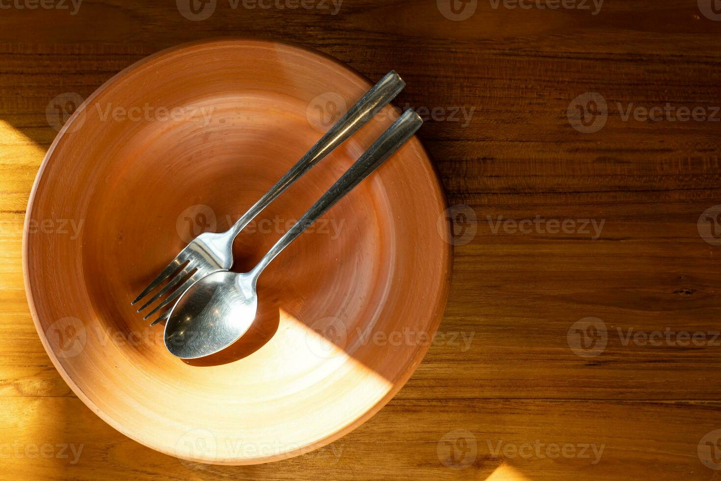 schließen oben rostfrei Stahl Besteck auf Lehm Platte. Lehm Teller mit Löffel und Gabel auf hölzern Tabelle mit Sonnenlicht. foto
