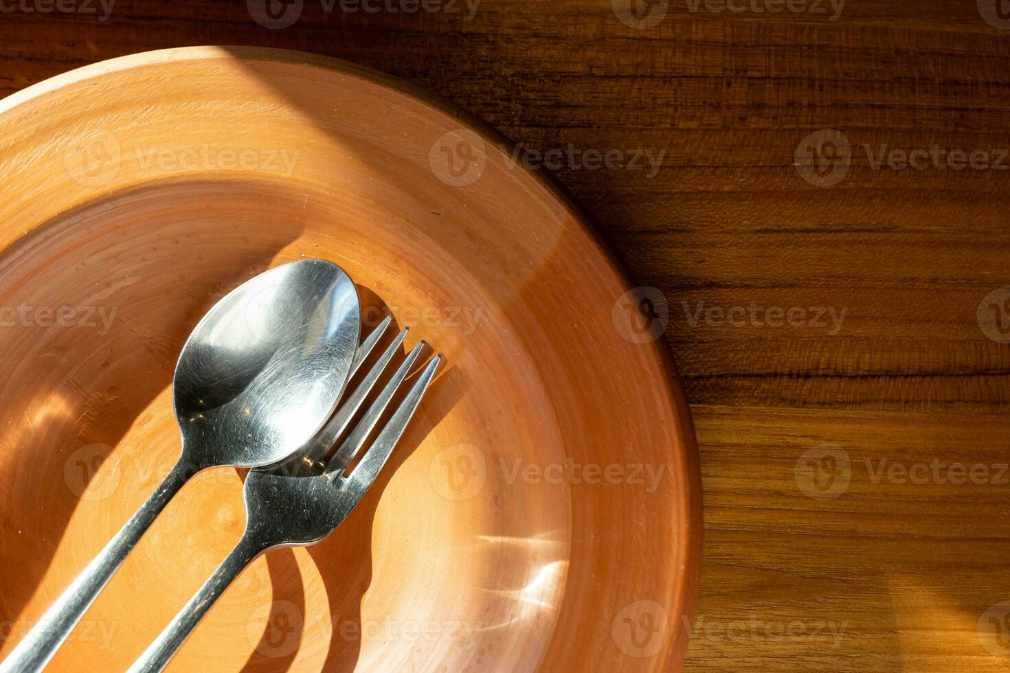 schließen oben rostfrei Stahl Besteck auf Lehm Platte. Lehm Teller mit Löffel und Gabel auf hölzern Tabelle mit Sonnenlicht. foto