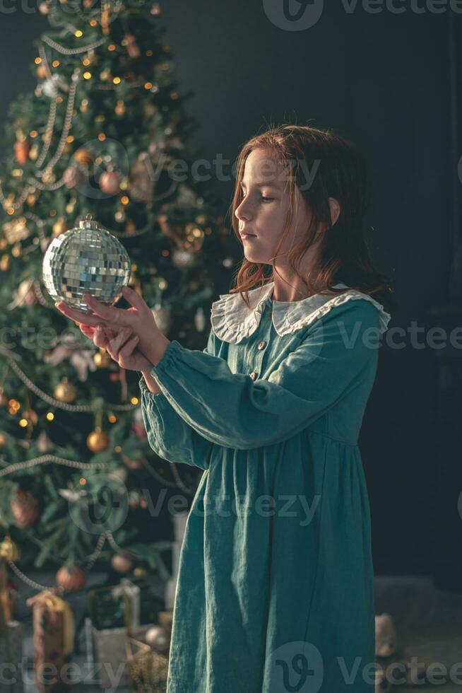 süß Mädchen hält ein Disko Ball gegen das Hintergrund von ein Weihnachten Baum foto