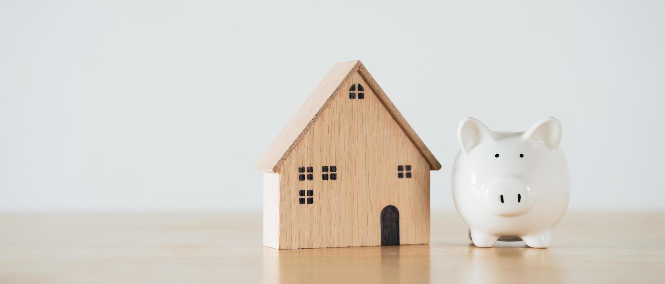 Holzhaus mit weißem Sparschwein auf Holztisch. Geld sparen für den Kauf eines Hauses, Finanzierungskonzept für Wohnungsbaudarlehen. foto
