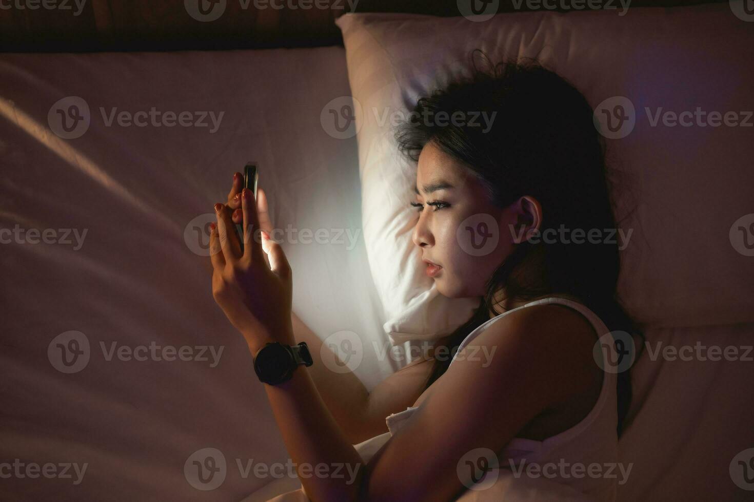 asiatisch Frau mit Handy, Mobiltelefon Telefon Smartphone Verlegung auf das Bett im das Schlafzimmer. schläfrig erschöpft, können nicht schlafen. Schlaflosigkeit, Sucht Konzept. Frauen Scrollen Sozial Netzwerke auf Handy, Mobiltelefon dunkel Schlafzimmer. foto
