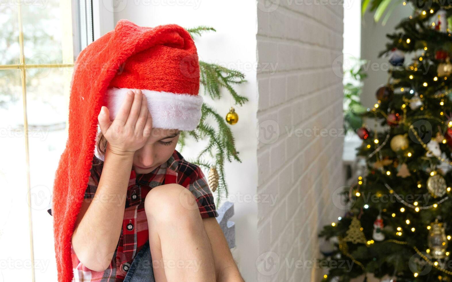 unglückliches mädchen in einer weihnachtsmütze sitzt auf der fensterbank des hauses in der nähe des weihnachtsbaums. das kind ist verärgert und weint und wartet auf weihnachten und neujahr. es gibt keine Geschenke, Freude und Wunder, Enttäuschung foto