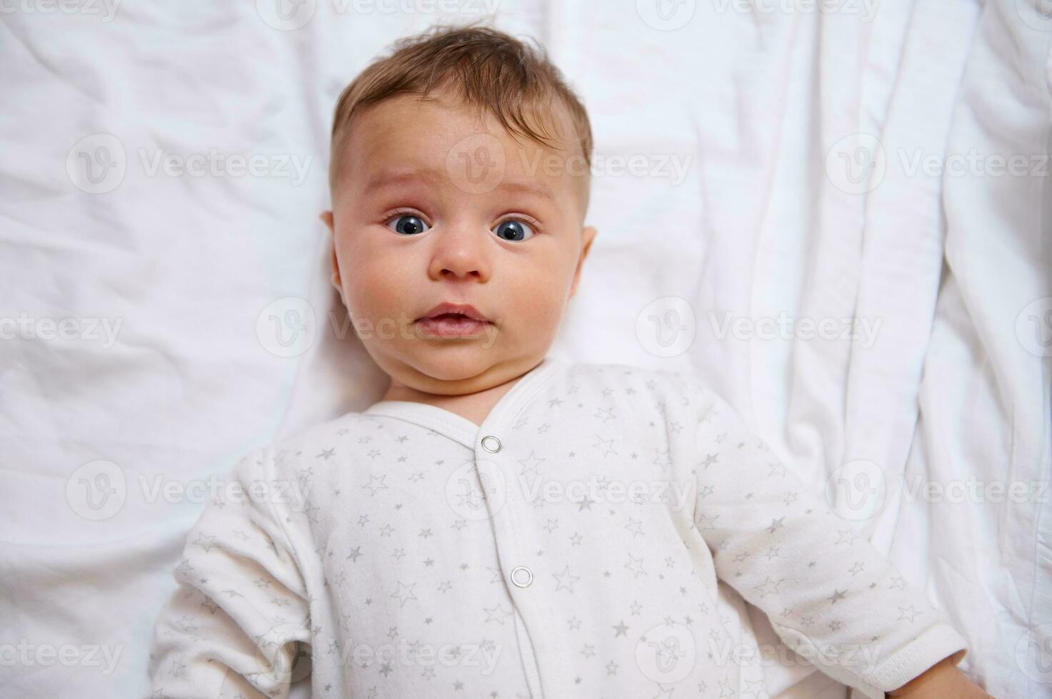 Nahansicht Porträt von süß Baby Junge 4-6 Monate alt suchen beim Kamera, Lügen auf Weiß Bett Blätter auf das Bett. Kopieren Anzeige Raum foto