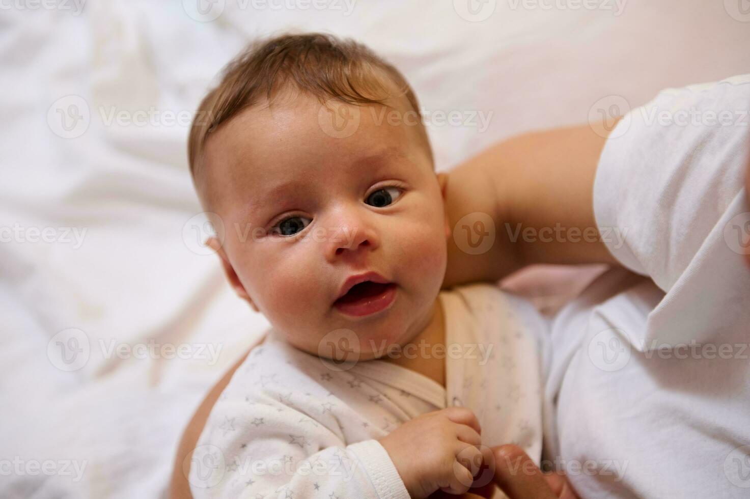 Nahansicht Porträt von bezaubernd süß schön Neugeborene Baby Junge im Weiß Kleidung, suchen beim Kamera während seine Mama kuschelt ihm foto