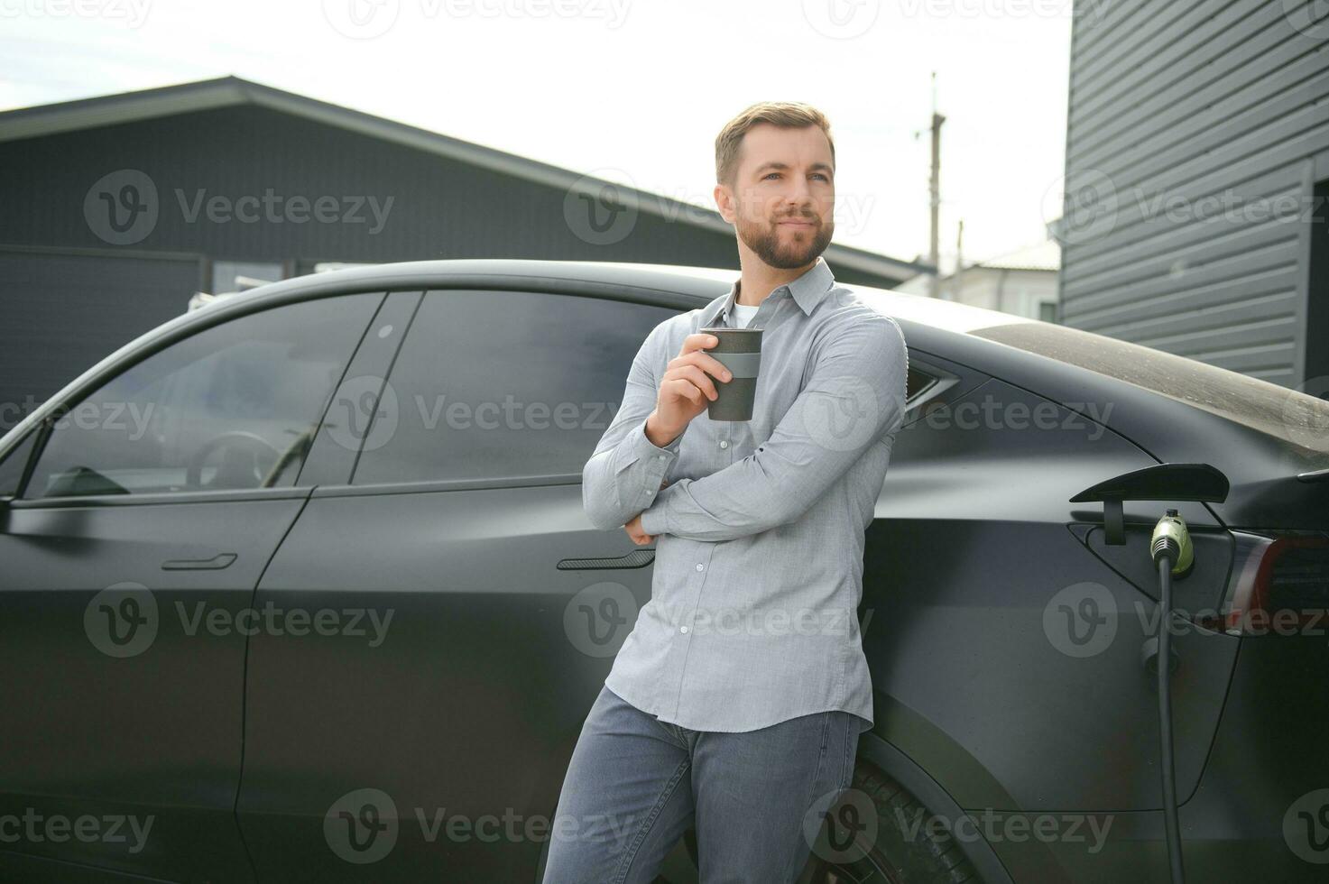 gut aussehend Mann Trinken Kaffee während Laden elektrisch Auto foto