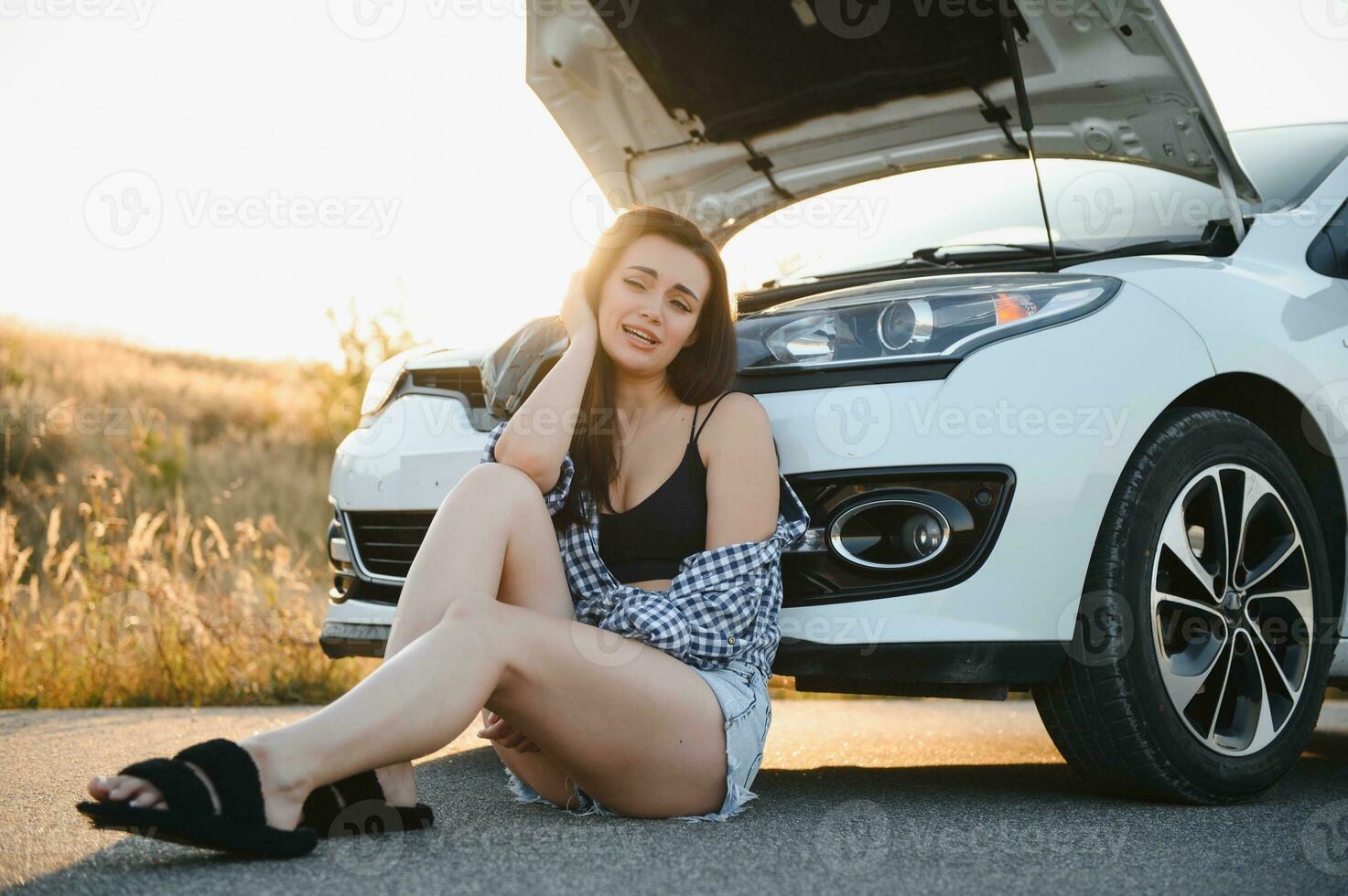 ein jung Mädchen sitzt in der Nähe von ein gebrochen Auto auf das Straße mit ein öffnen Haube. foto