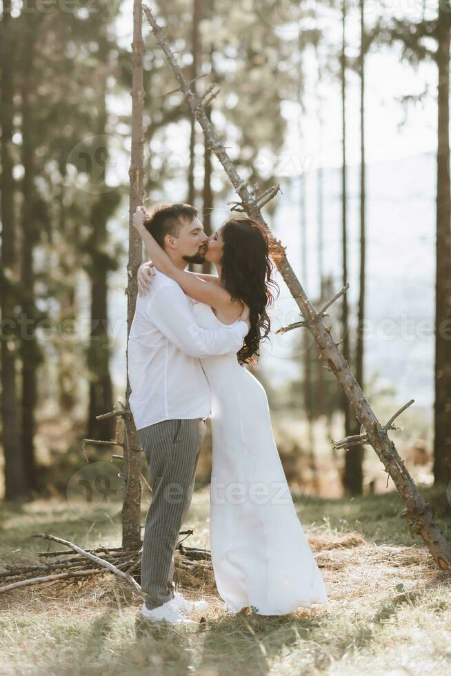 stilvoll Bräutigam im Weiß Hemd und süß Brünette Braut im Weiß Kleid im Wald in der Nähe von Hochzeit hölzern Bogen. Hochzeit Porträt von Jungvermählten. foto