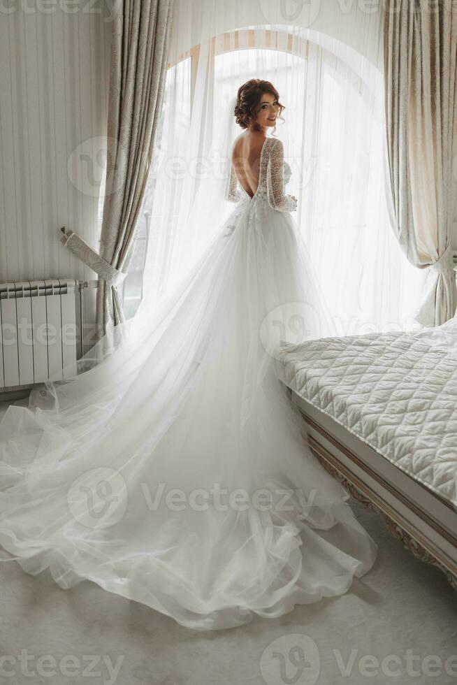 das Braut im ein schön Kleid ist Stehen durch das Fenster mit ein lange Zug. Hochzeit. Hochzeit Strauß Morgen von das Braut. ein glücklich Braut ist warten zum das Bräutigam. foto