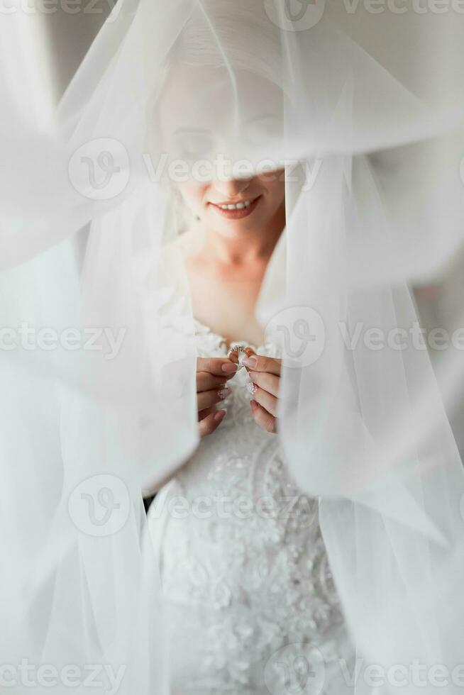 ein blond Braut im ein Weiß Kleid ist halten ihr Gold Hochzeit Ring. Französisch Maniküre. öffnen Schultern. schön Hände. lange Schleier. Morgen von das Braut. Einzelheiten foto