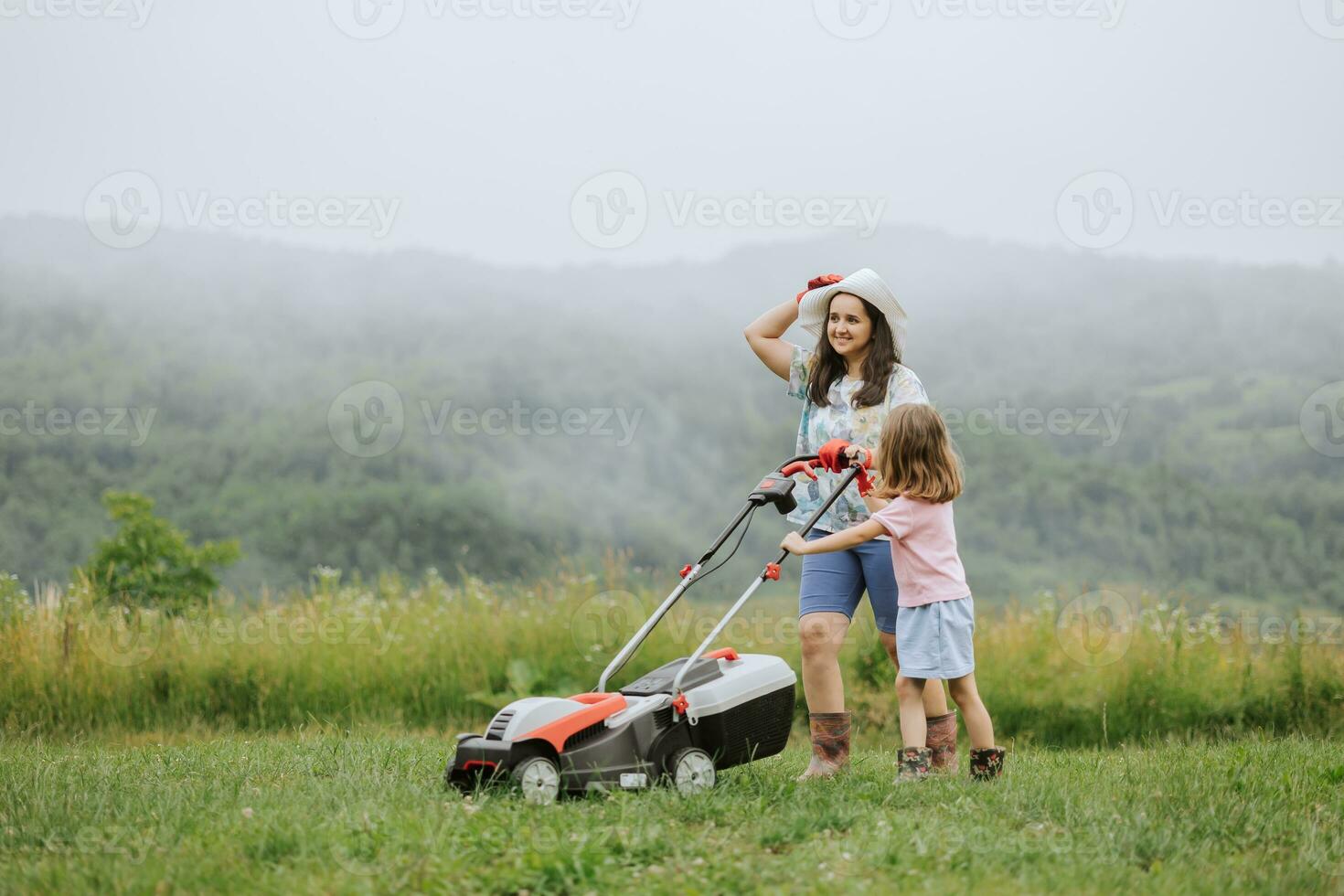 ein Frau im Stiefel mit ihr Kind im das bilden von ein Spiel mäht das Gras mit ein Rasenmäher im das Garten gegen das Hintergrund von Berge und Nebel, Garten Werkzeuge Konzept, arbeiten, Natur. foto