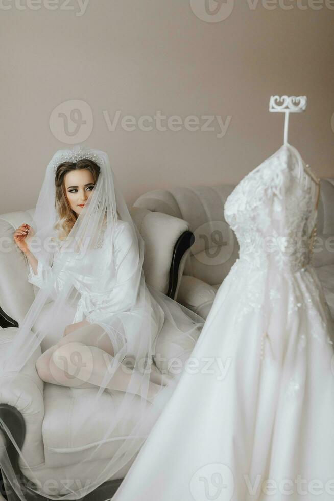 das blond Braut ist gekleidet im ein Satin- Kleid von Sitze und ein Sofa unter ein Schleier. schön Haar und bilden, öffnen Büste. Hochzeit Porträt. ein schön Braut im ein Dressing Kleid posiert Nächster zu ihr Kleid foto