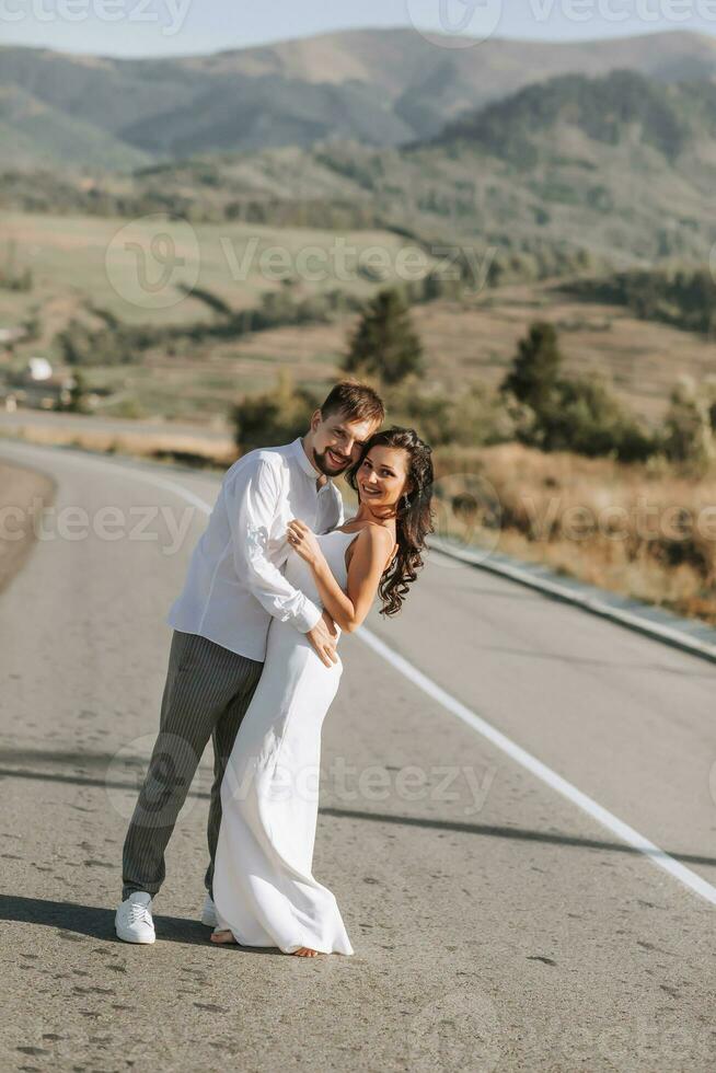 ein stilvoll Bräutigam im ein Weiß Hemd und ein süß Brünette Braut im ein Weiß Kleid sind umarmen und suchen beim das Kamera auf ein Asphalt Straße gegen das Hintergrund von ein Wald und Berge. foto