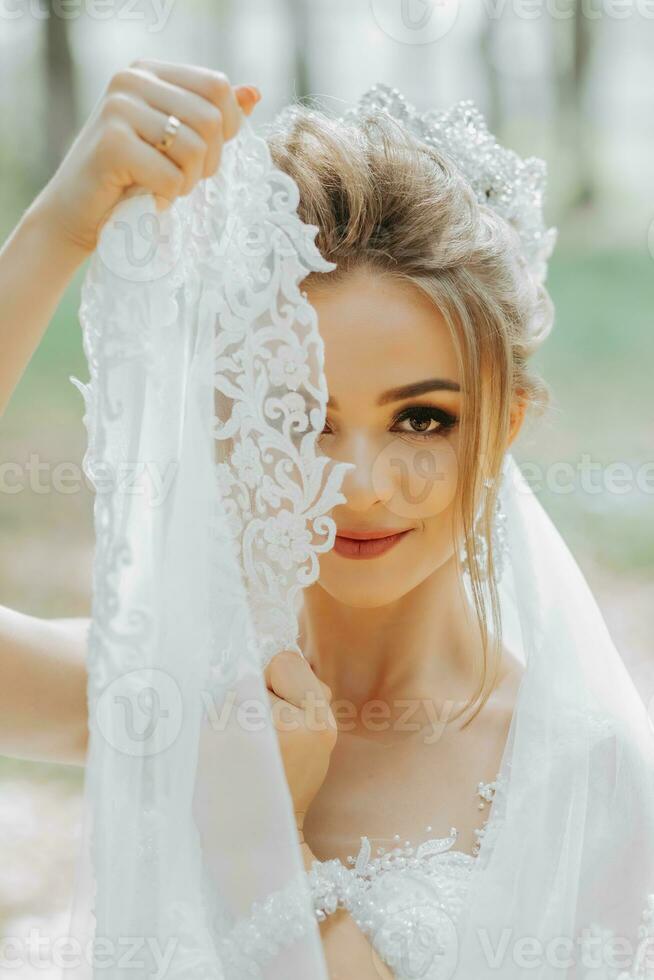 schön jung Frau im ein Hochzeit Kleid im das Wald mit ein königlich Frisur und ein schick Tiara hält ihr Schleier im ihr Hände, Hochzeit im Gold Farbe foto