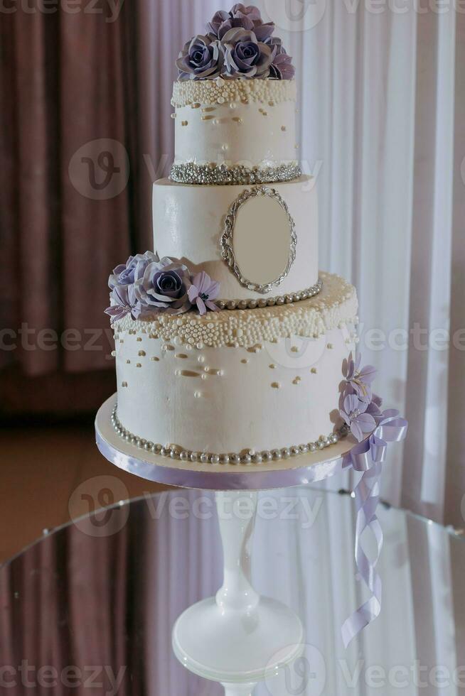 Weiß dreistufig Hochzeit Kuchen dekoriert mit lila Blumen und Perlen, Stehen auf ein Spiegel Stand. Nachspeisen Hochzeit. foto