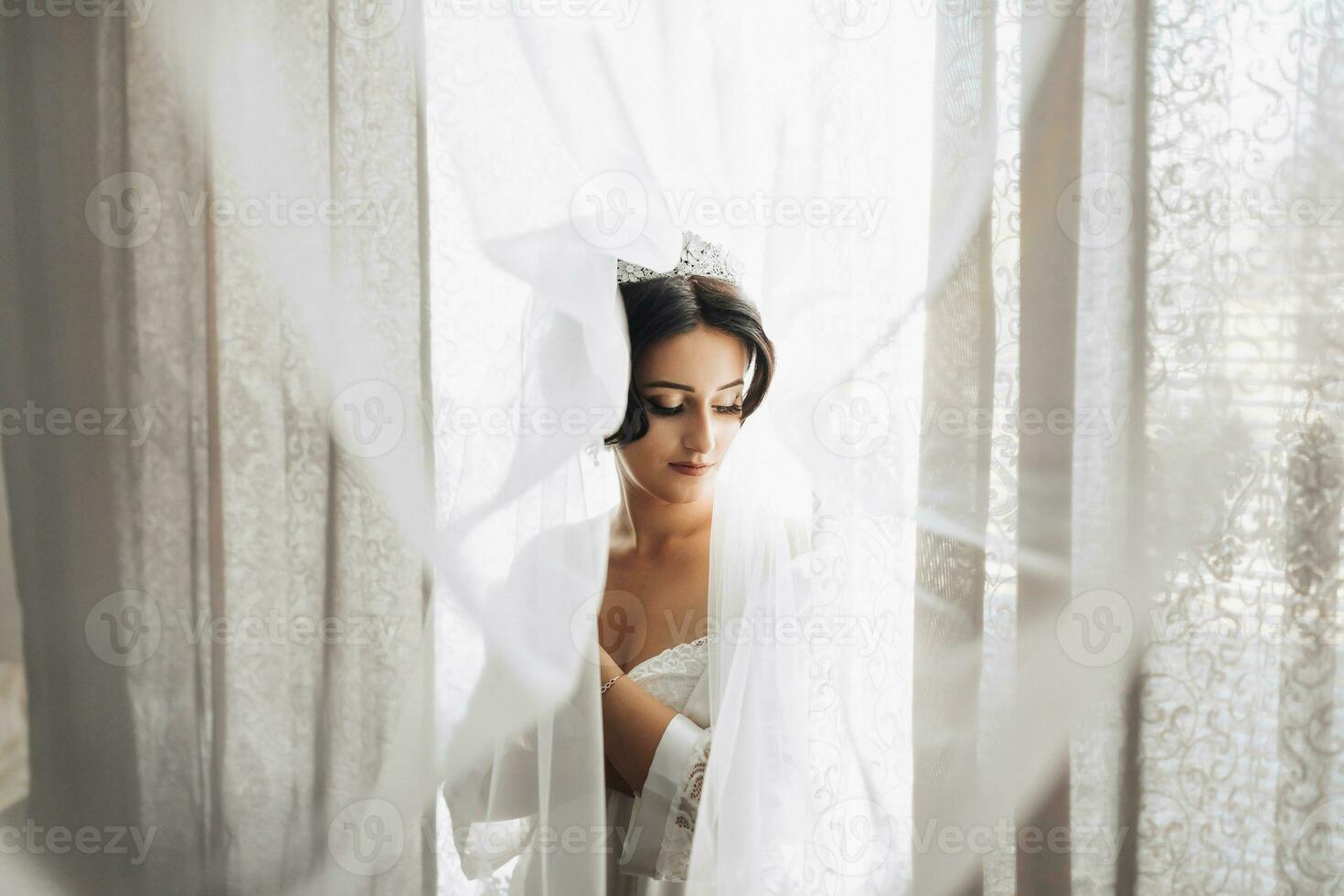 Porträt. ein Brünette Braut ist Stehen eingewickelt im ein Schleier durch das Fenster und suchen runter. Herrlich bilden und Haar. voluminös Schleier. Hochzeit Foto. schön Braut foto