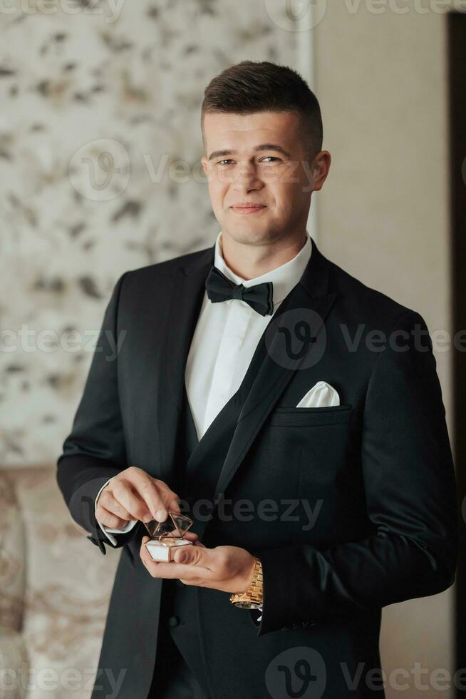 Porträt. ein Mann im ein Weiß Shirt, ein schwarz Bogen Krawatte und ein schwarz passen posiert im ein Zimmer mit Hochzeit Schuhe. ein stilvoll betrachten. Herren Stil. Mode. Geschäft foto