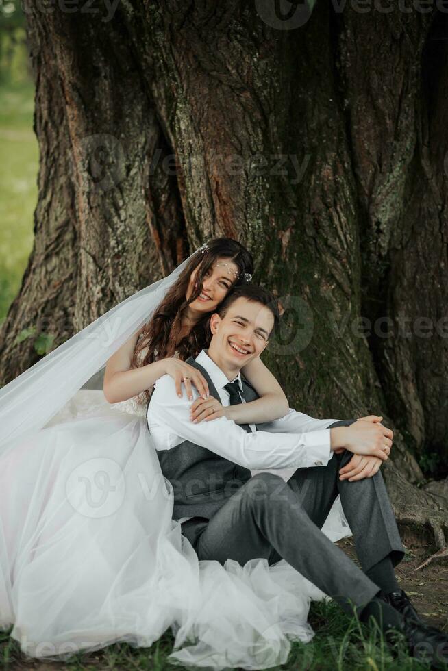 ein schön Braut mit lange lockig Haar im ein schick Kleid Umarmungen das Bräutigam, lächelt, suchen in das Linse unter ein groß Baum. Porträt von das Braut und Bräutigam. Frühling Hochzeit. natürlich bilden foto