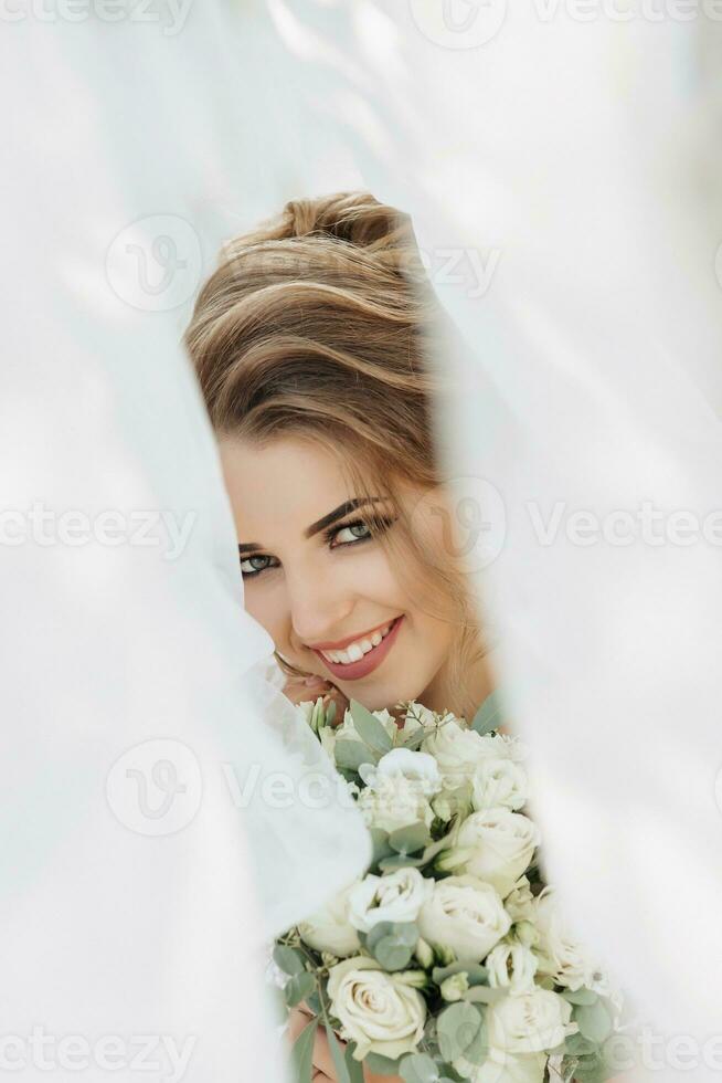 lockig blond Braut im ein Weiß Kleid, bedeckt mit ein Schleier, posiert zum das Kamera mit ein Strauß von Rosen. Porträt von das Braut. schön bilden und Haar. Hochzeit im Natur foto