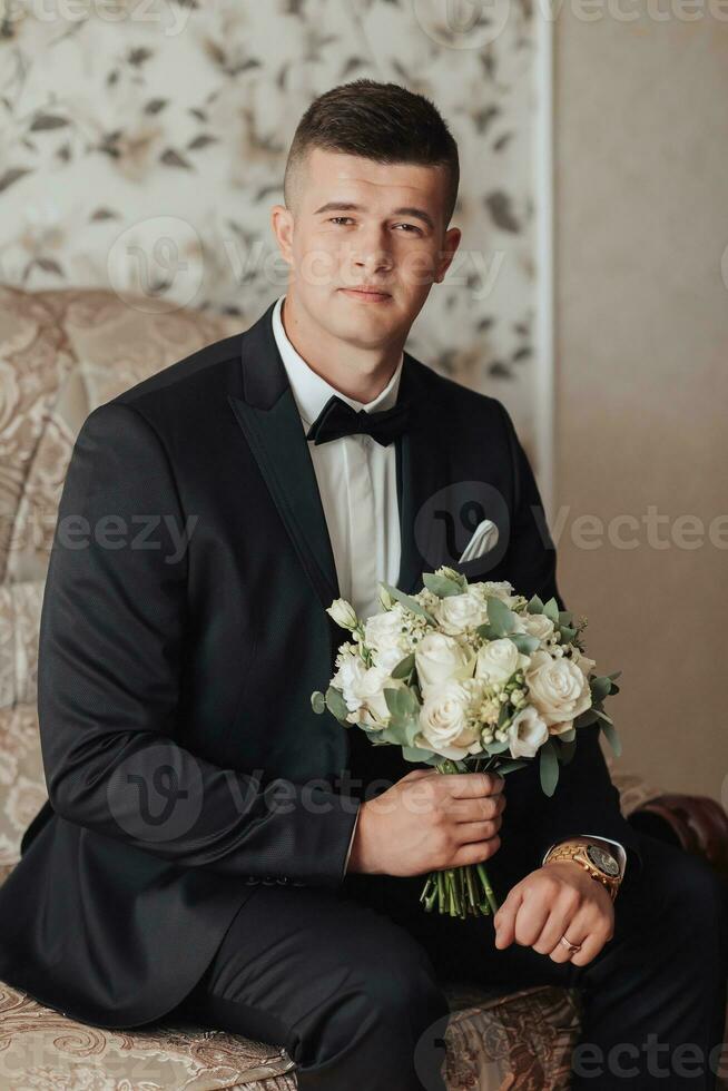 Porträt. ein Mann im ein Weiß Shirt, ein schwarz Bogen Krawatte und ein schwarz passen posiert im ein Zimmer mit ein Hochzeit Strauß. ein stilvoll betrachten. Herren Stil. Mode. Geschäft foto