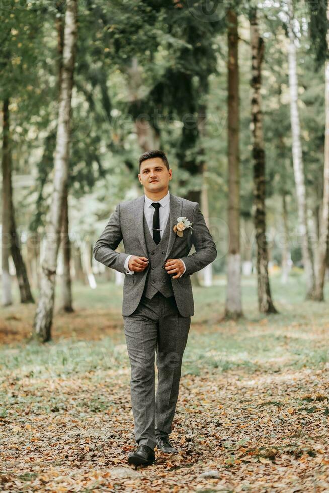 ein stilvoll Bräutigam Spaziergänge nach vorne im das Wald Knöpfen seine Jacke zwischen hoch Bäume. ein modisch Bräutigam, ein Erwachsene Mann foto