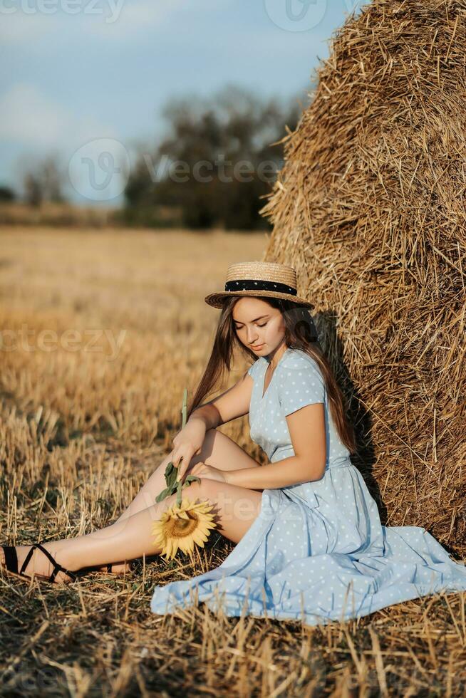 Porträt von ein jung Mädchen. ein Mädchen im ein Blau Kleid und Hut, halten ein Strauß von Sonnenblumen, gegen ein Hintergrund von Heu Ballen. lange Gerade Haar. nett Farbe. Sommer- foto