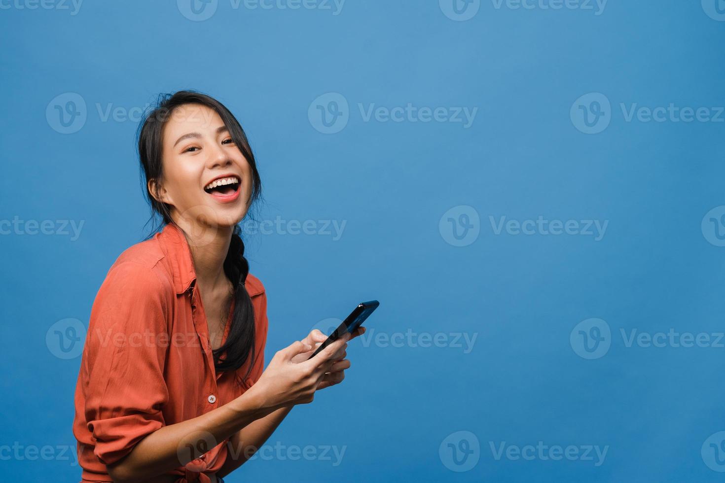 überraschte junge asiatische dame, die handy mit positivem ausdruck benutzt, breit lächelt, in lässiger kleidung gekleidet ist und die kamera auf blauem hintergrund betrachtet. glückliche entzückende frohe frau freut sich über erfolg. foto