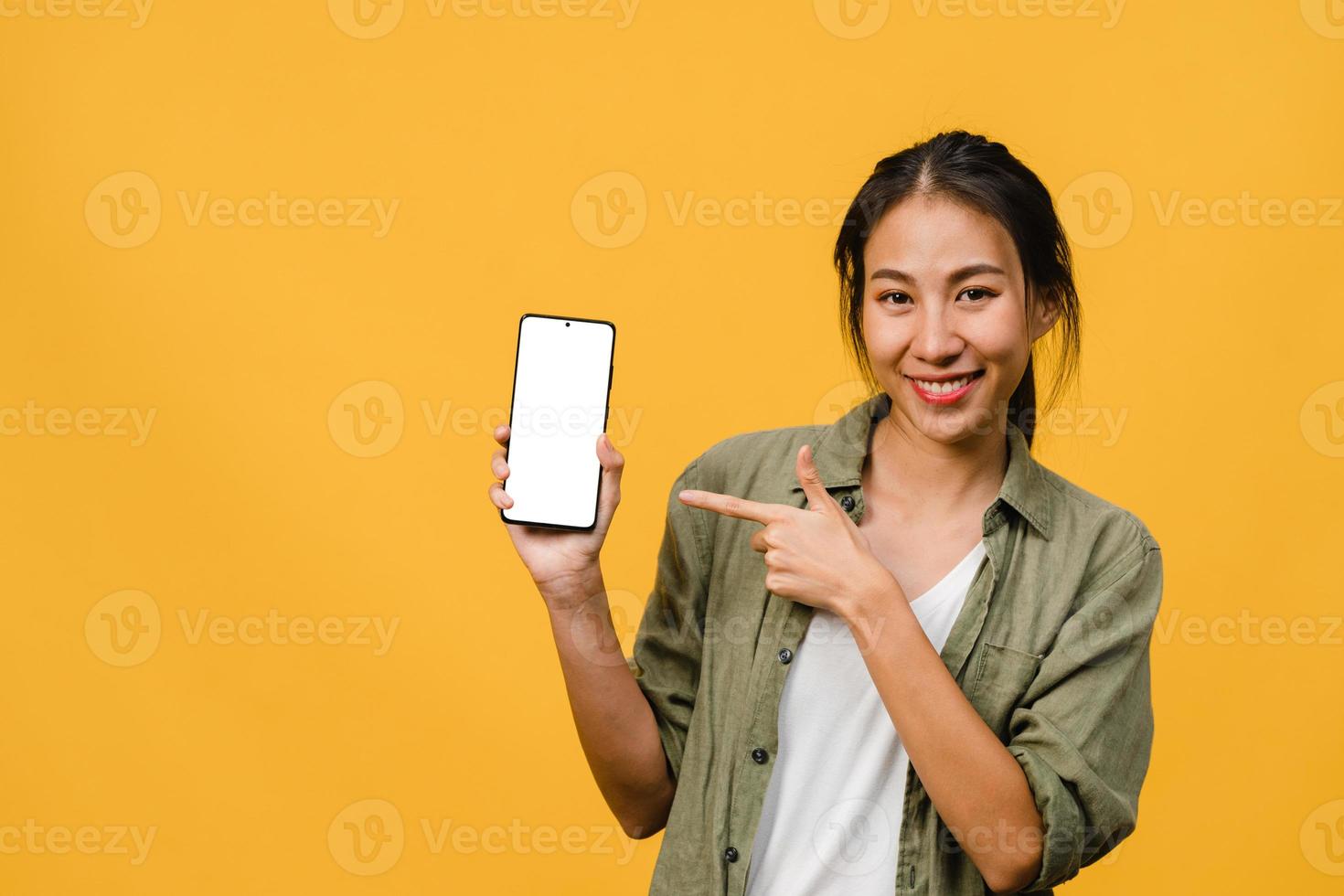 junge asiatische Dame zeigt leeren Smartphone-Bildschirm mit positivem Ausdruck, lächelt breit, gekleidet in Freizeitkleidung und fühlt sich glücklich auf gelbem Hintergrund. Handy mit weißem Bildschirm in weiblicher Hand. foto