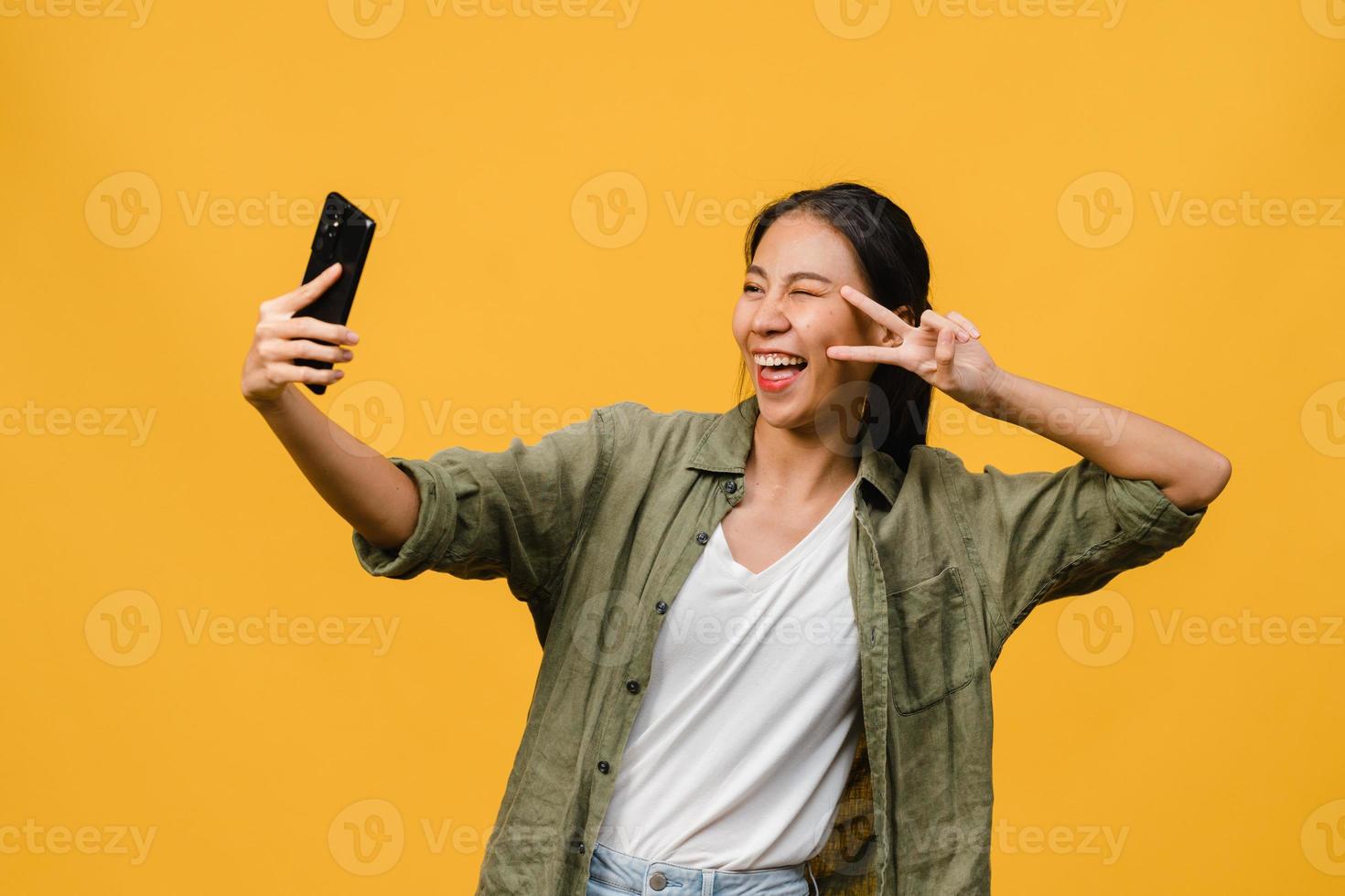 Lächelnde entzückende asiatische Frau, die Selfie-Foto auf Smartphone mit positivem Ausdruck in Freizeitkleidung macht und auf gelbem Hintergrund steht. glückliche entzückende frohe frau freut sich über erfolg. foto