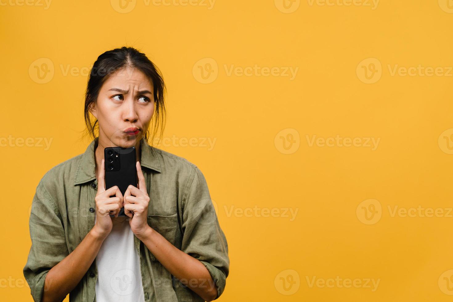 Denken träumende junge asiatische Dame mit Telefon mit positivem Ausdruck, gekleidet in lässiges Tuch, das Glück fühlt und isoliert auf gelbem Hintergrund steht. glückliche entzückende frohe frau freut sich über erfolg. foto