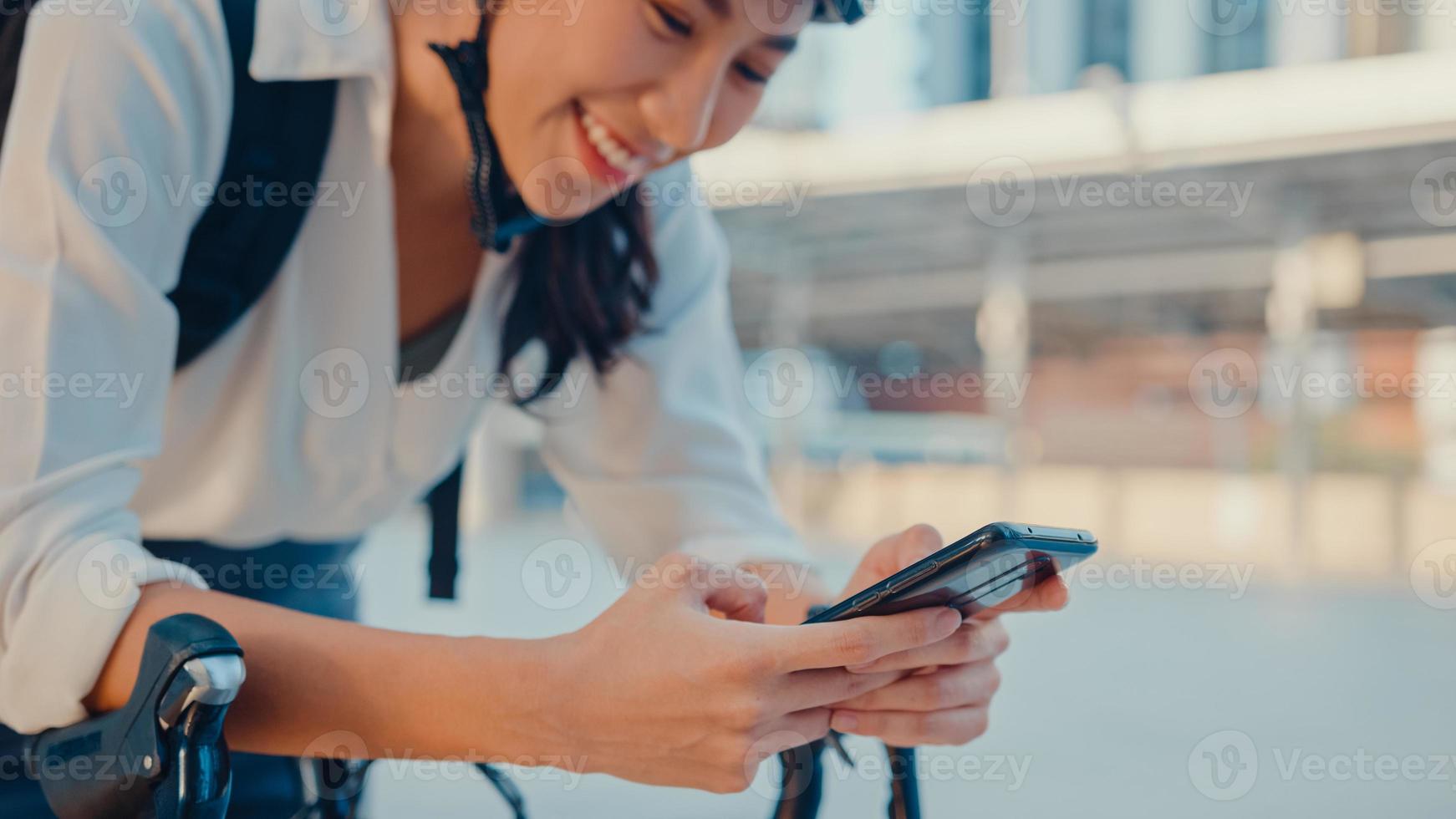 Lächeln Sie asiatische Geschäftsfrau mit Rucksack, die eine Smartphone-Look-Kamera im Stadtstand an der Straße mit dem Fahrrad verwendet, um im Büro zu arbeiten. Sportmädchen benutzen Telefon für die Arbeit. zur Arbeit pendeln, Berufspendler in der Stadt. foto