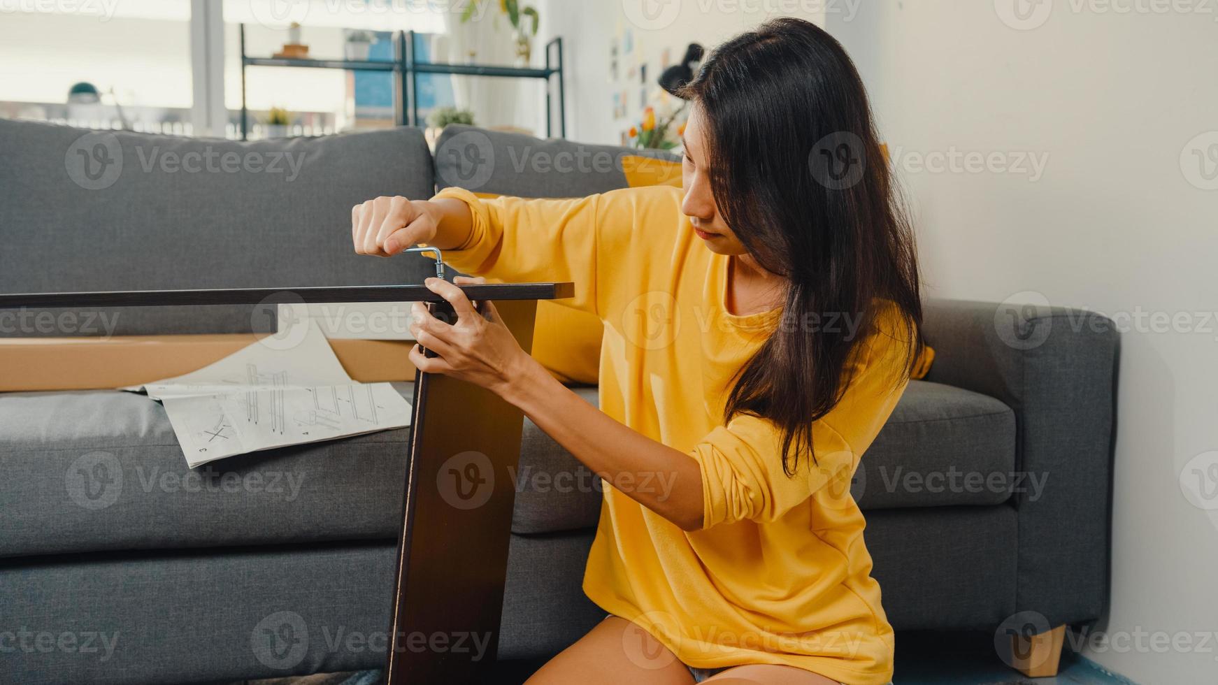 glückliche asiatische junge frau, die schachtel auspackt und die anweisungen zum zusammenbauen neuer möbel liest, dekorieren hausbautisch mit karton im wohnzimmer zu hause. foto