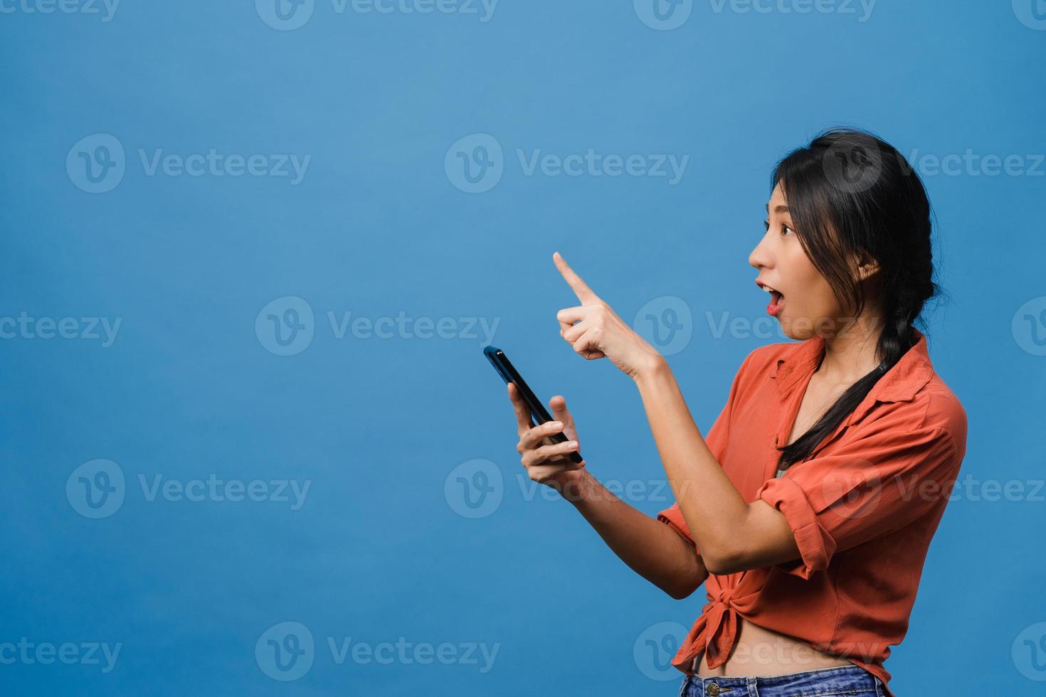 Porträt einer jungen asiatischen Dame, die Handy mit fröhlichem Ausdruck verwendet, etwas Erstaunliches an Leerzeichen in Freizeitkleidung zeigt und auf blauem Hintergrund isoliert steht. Gesichtsausdruck Konzept. foto