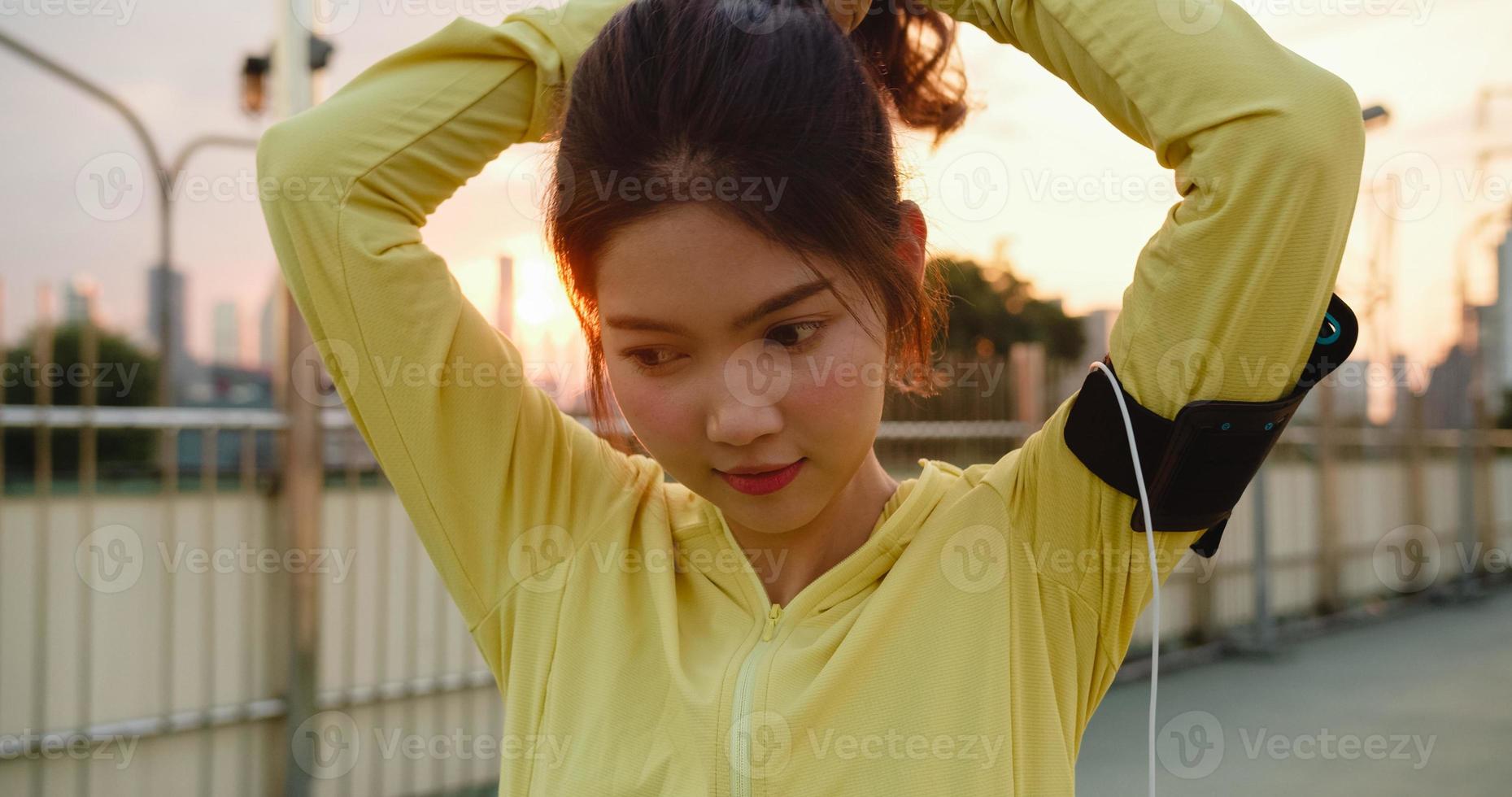 glückliche junge asiatische Sportlerin in gelber Kleidung, die sich auf das Training im städtischen Umfeld vorbereitet. Koreanisches Teenie-Mädchen, das sich Pferdeschwanzhaare bindet und sich vor ihrem Training auf der Gehwegbrücke am frühen Morgen fertig macht foto
