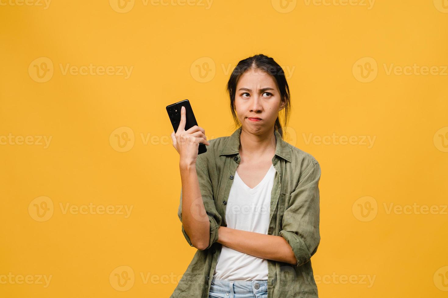 Denken träumende junge asiatische Dame mit Telefon mit positivem Ausdruck, gekleidet in lässiges Tuch, das Glück fühlt und isoliert auf gelbem Hintergrund steht. glückliche entzückende frohe frau freut sich über erfolg. foto