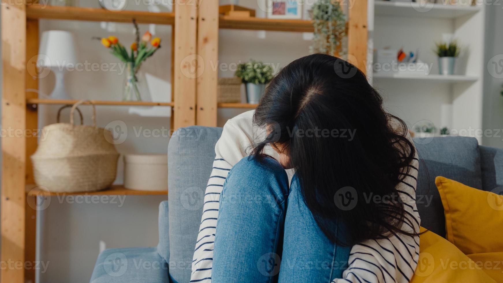 nachdenkliche asiatische dame sitzt auf dem sofa im wohnzimmer im haus auf den knien und schaut nach draußen mit dem gefühl, einsam zu sein, traurige depressive teenager verbringen zeit allein zu hause, soziale distanz, coronavirus-quarantäne. foto