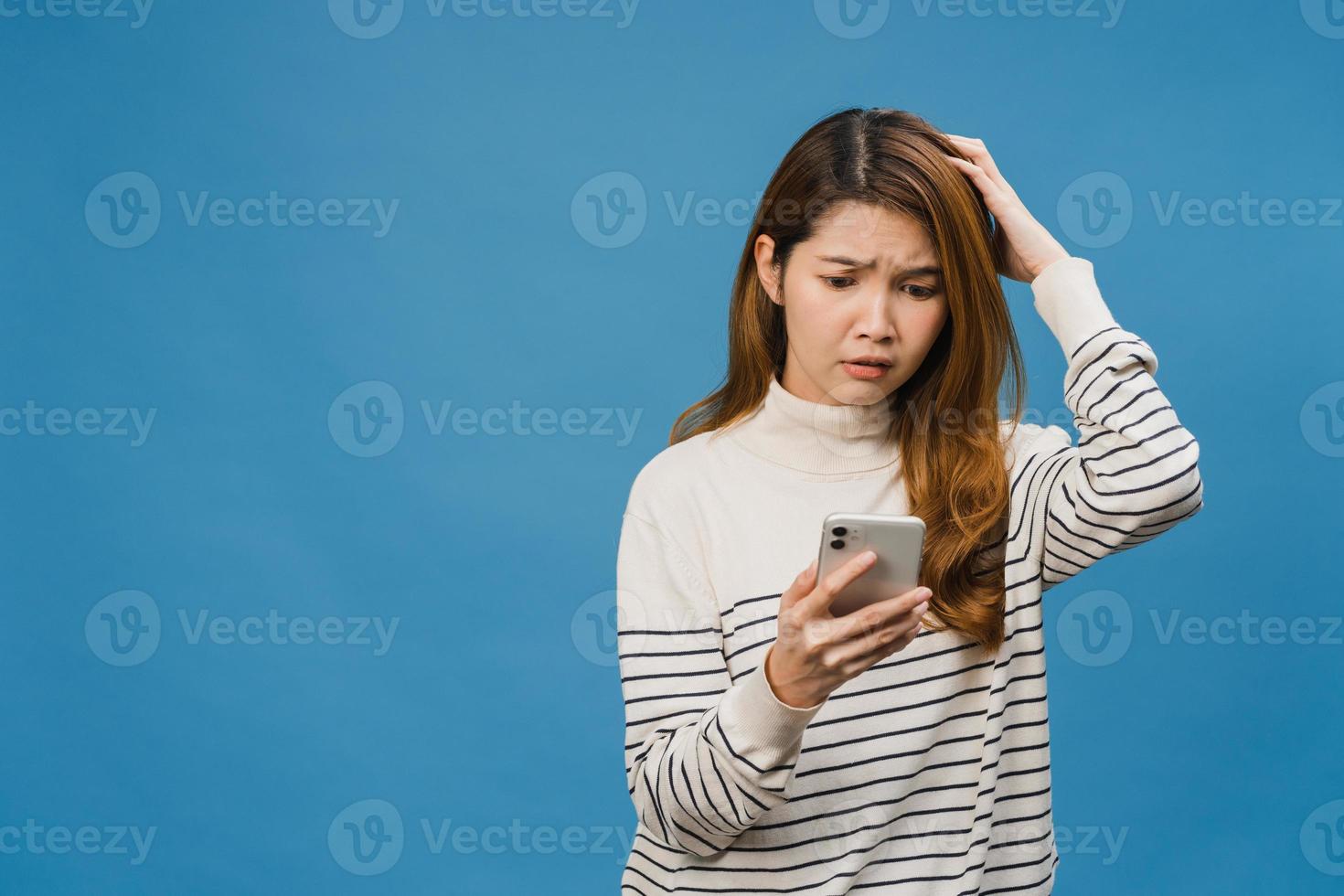 Denken träumende junge asiatische Dame mit Telefon mit positivem Ausdruck, gekleidet in Freizeitkleidung, die sich glücklich fühlt und isoliert auf blauem Hintergrund steht. glückliche entzückende frohe frau freut sich über erfolg. foto