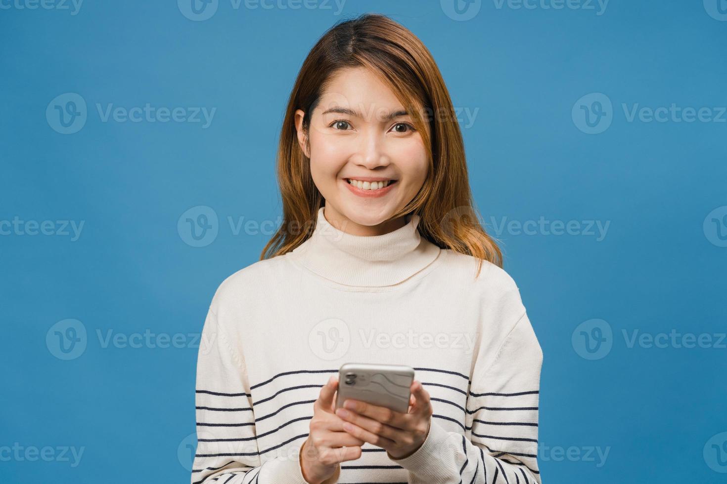 überraschte junge asiatische dame, die handy mit positivem ausdruck benutzt, breit lächelt, in lässiger kleidung gekleidet ist und die kamera auf blauem hintergrund betrachtet. glückliche entzückende frohe frau freut sich über erfolg. foto