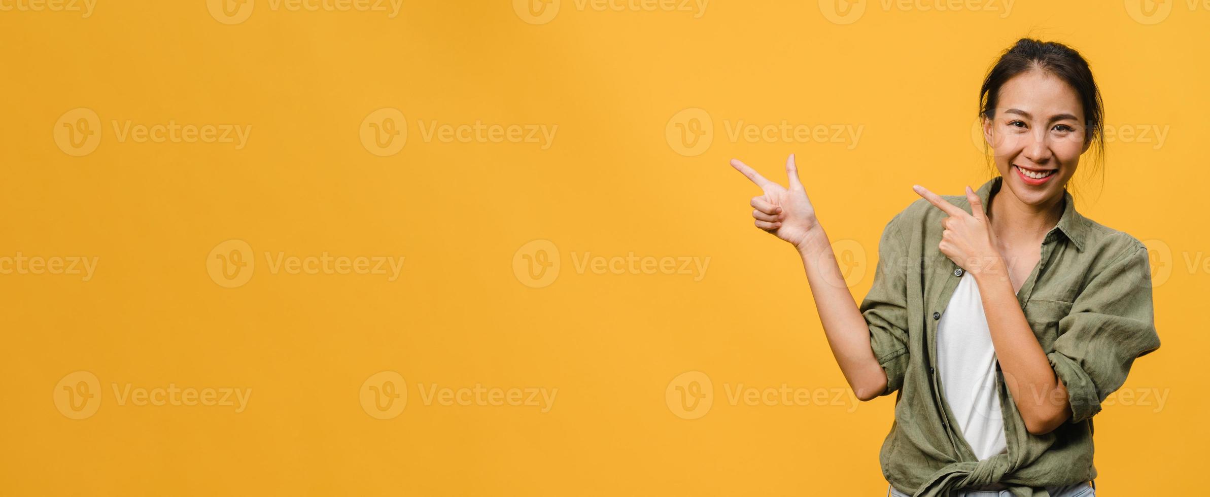 junge asiatische dame, die mit fröhlichem ausdruck lächelt, zeigt etwas erstaunliches an leeren räumen in lässigem tuch und blickt auf die kamera einzeln auf gelbem hintergrund. Panoramabanner mit Kopienraum. foto
