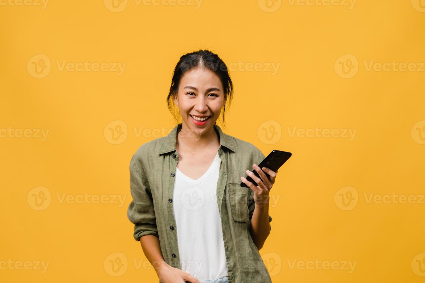 überraschte junge asiatische dame, die handy mit positivem ausdruck benutzt, breit lächelt, in lässiger kleidung gekleidet ist und die kamera auf gelbem hintergrund betrachtet. glückliche entzückende frohe frau freut sich über erfolg. foto