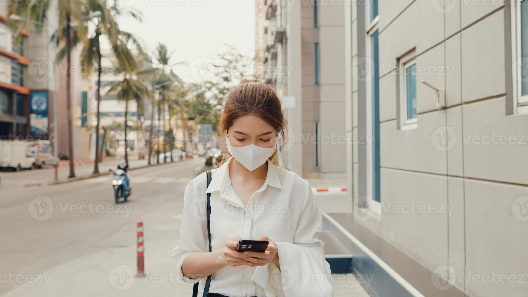 Erfolgreiche junge asiatische Geschäftsfrau in modischer Bürokleidung mit medizinischer Gesichtsmaske mit Smartphone, während sie morgens allein im Freien in der urbanen modernen Stadt spaziert. Business-on-Go-Konzept. foto