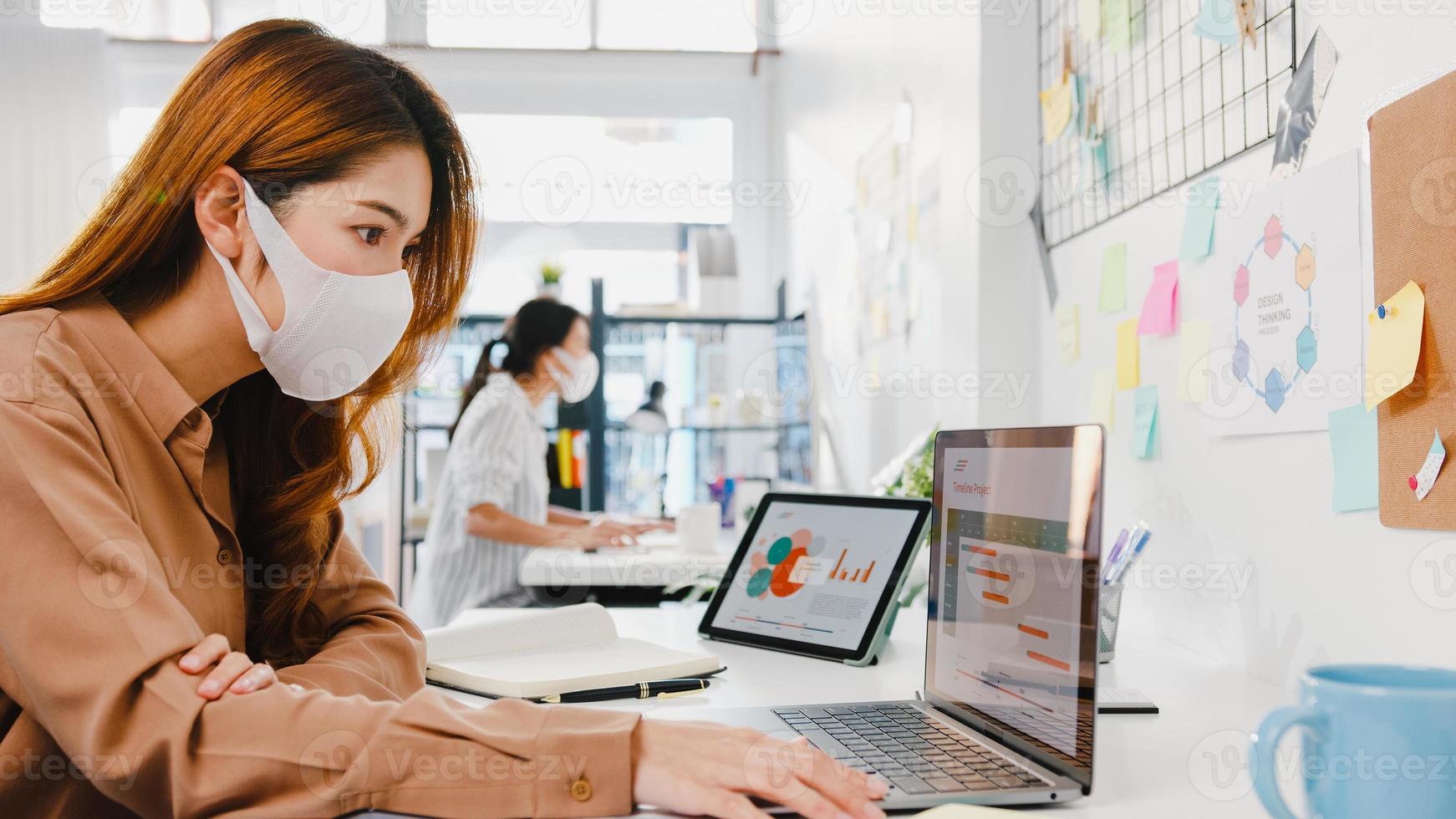 Asiatische Geschäftsfrau, die eine medizinische Gesichtsmaske für soziale Distanzierung in einer neuen normalen Situation zur Virusprävention trägt, während sie den Laptop wieder bei der Arbeit im Büro verwendet. Lebensstil nach dem Corona-Virus. foto