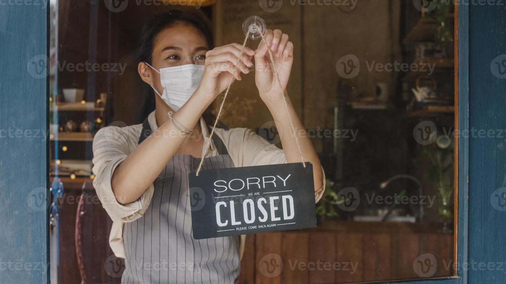 junge asiatische mädchen tragen gesichtsmaske, die ein zeichen von geschlossen zu offenem schild an der tür dreht und nach außen auf kunden wartet, nachdem sie gesperrt wurde. Inhaber Kleinunternehmen, Essen und Trinken, Geschäftskonzept wiedereröffnet. foto