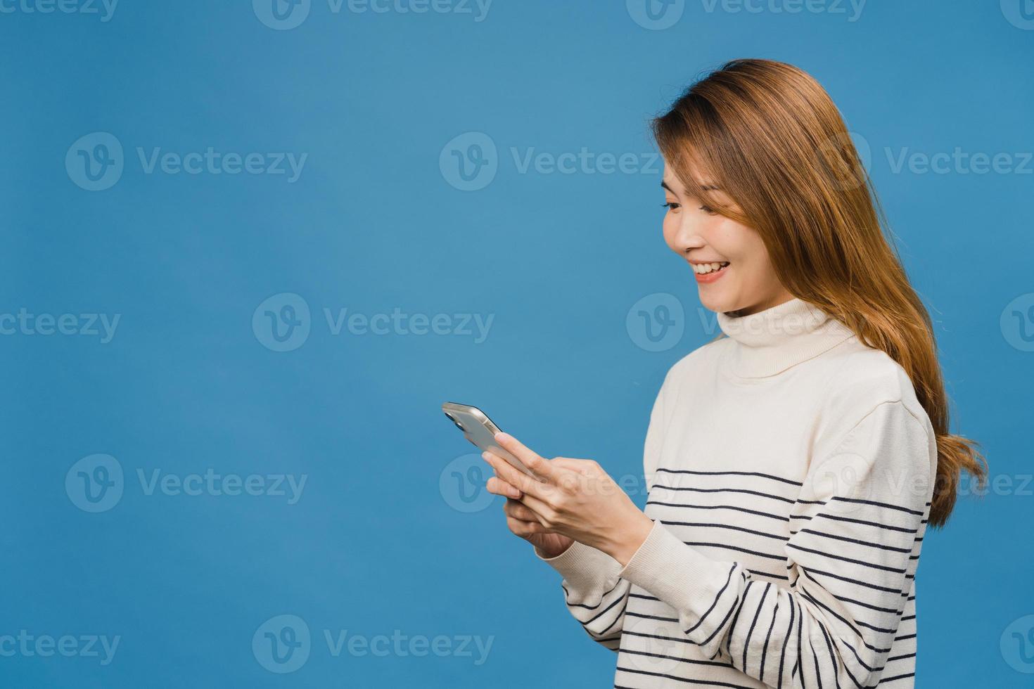 junge asiatische dame, die telefon mit positivem ausdruck verwendet, breit lächelt, in lässiger kleidung gekleidet, glücklich fühlt und isoliert auf blauem hintergrund steht. glückliche entzückende frohe frau freut sich über erfolg. foto