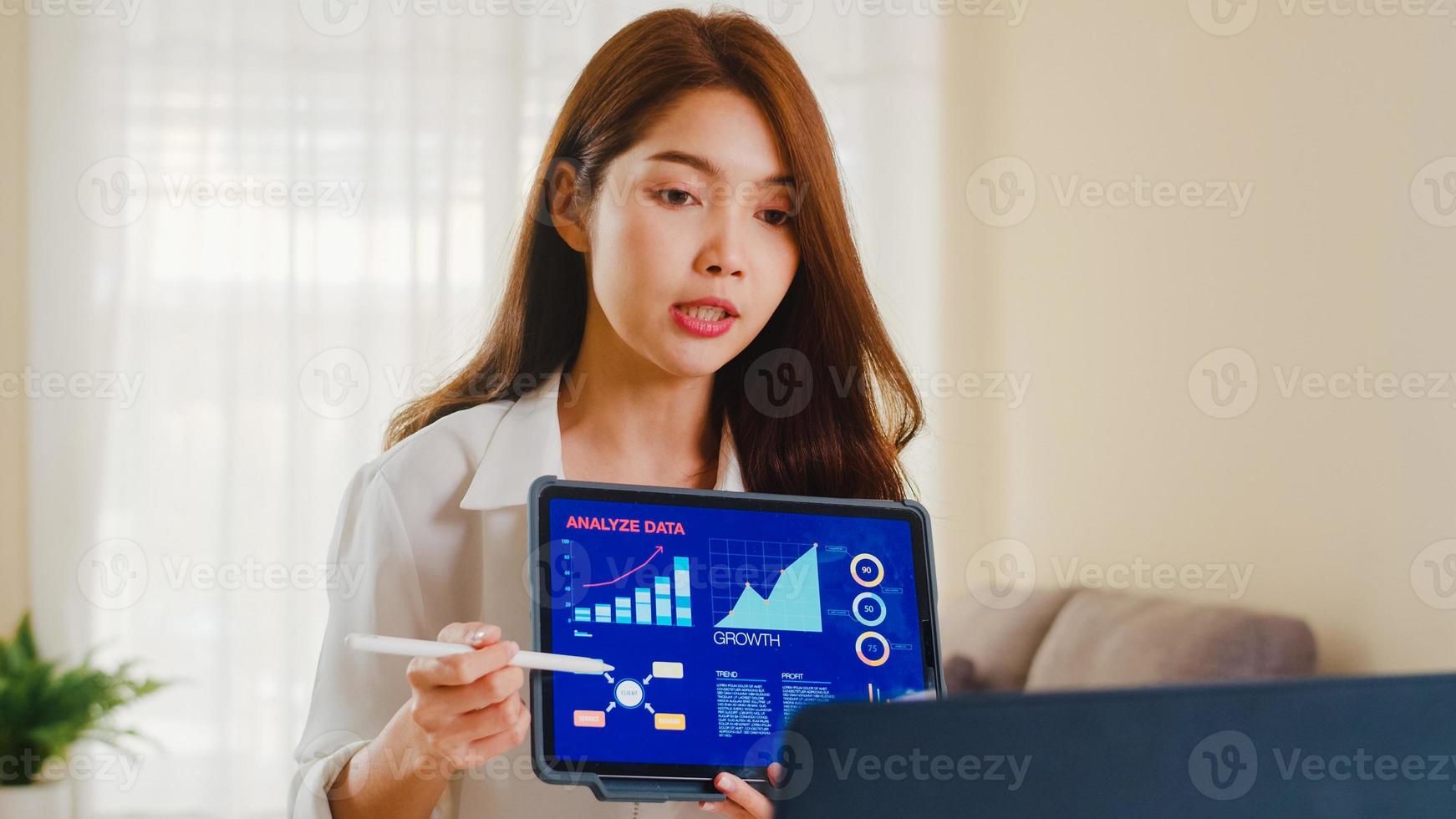 Asiatische Geschäftsfrau, die Laptop- und Tablet-Präsentation für Kollegen über den Plan im Videoanruf verwendet, während sie von zu Hause aus im Wohnzimmer arbeitet. Selbstisolation, soziale Distanzierung, Quarantäne für Coronavirus. foto